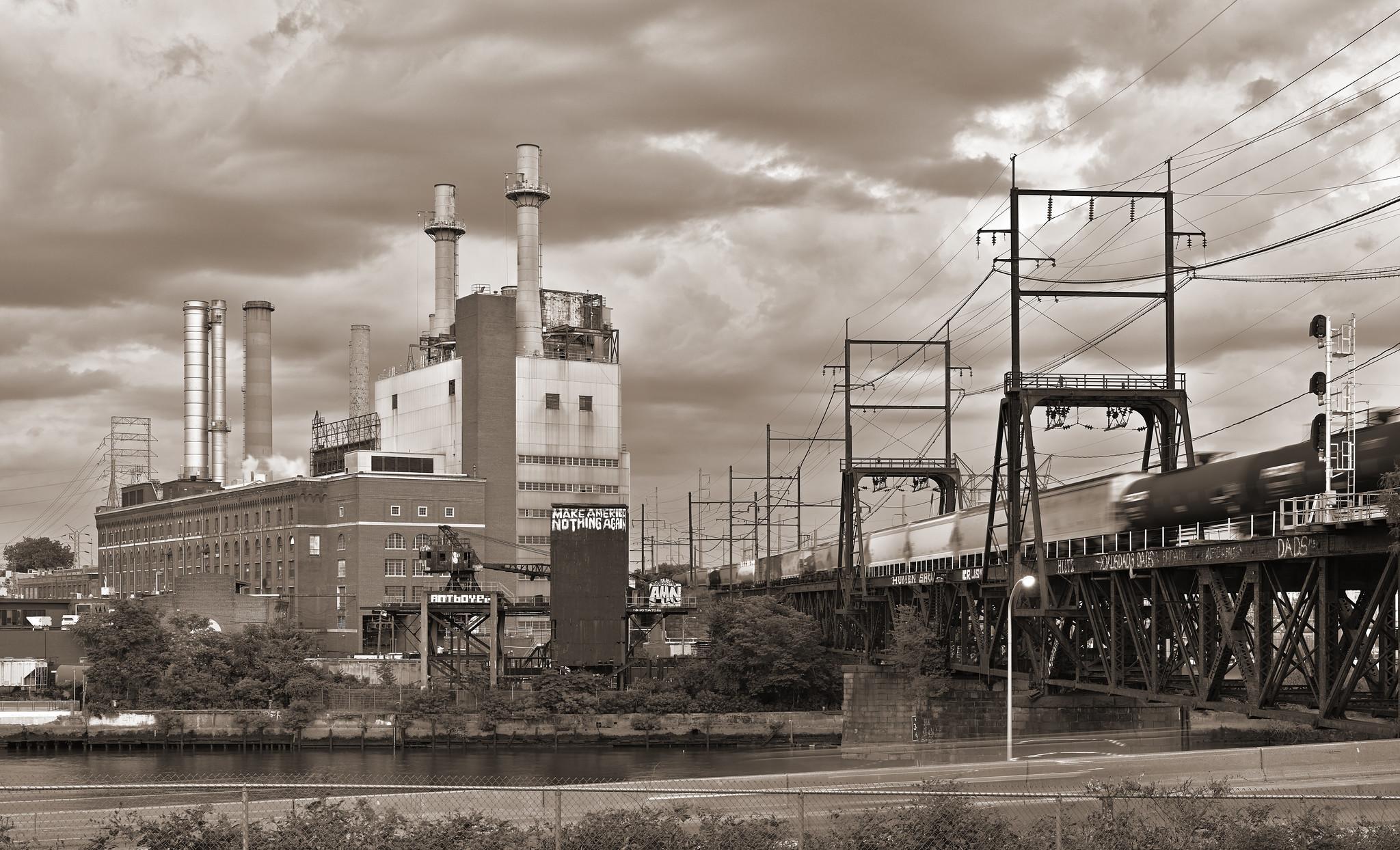 A mid-century power plant, with four prominent smokestacks, at left across a small river. At right, a railroad drawbridge crosses the river, with a freight train slowly streaking across.