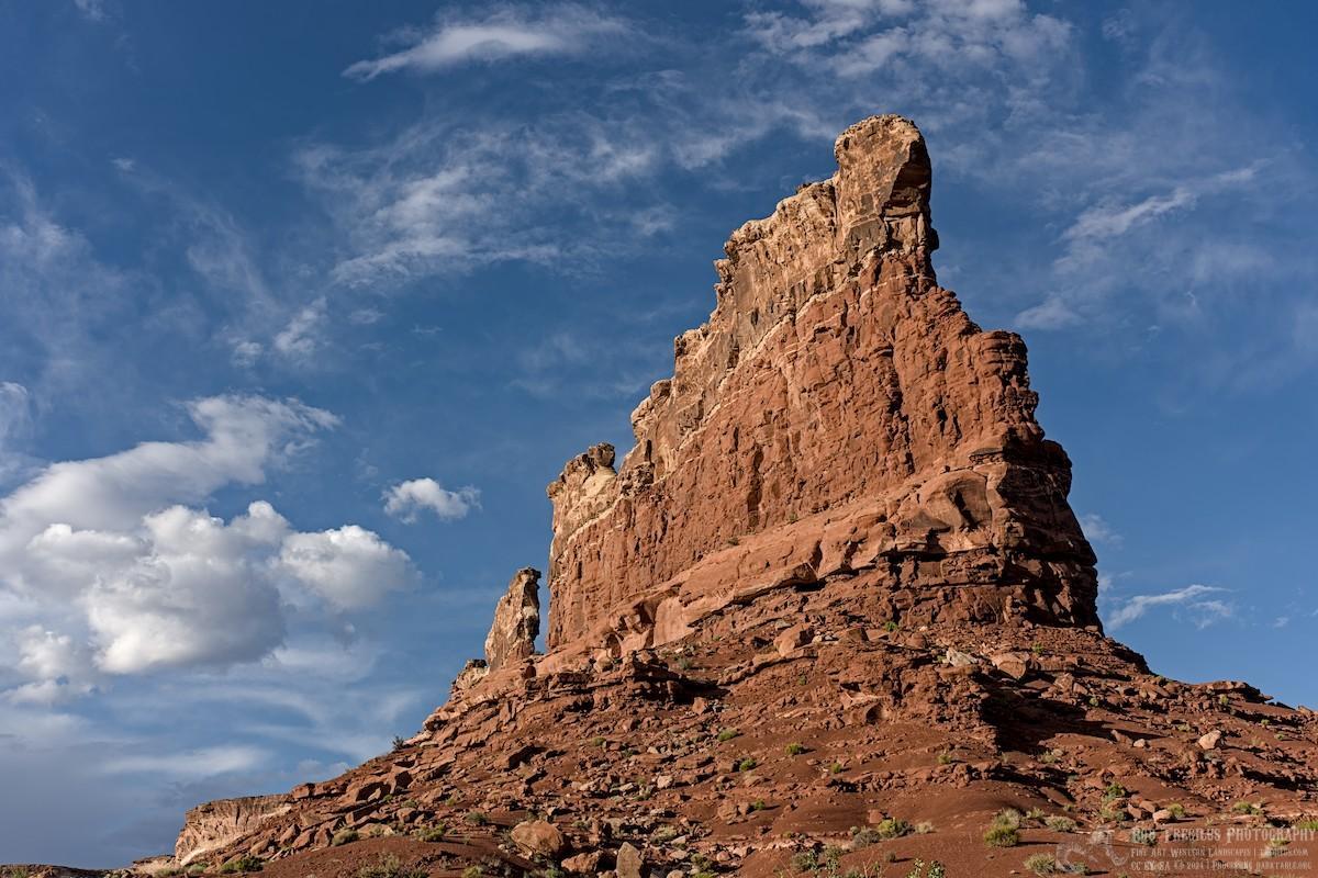 A color landscape photo of a very tall brown-red sandstone feature. It appears as a thin fin structure from center right and going toward center left. The view is from near the base looking up so the angle is dramatic. The fin is flat on top with a couple of missing sections, like a bad set of teeth. The sky is clear and very blue with wispy and puffy white clouds.