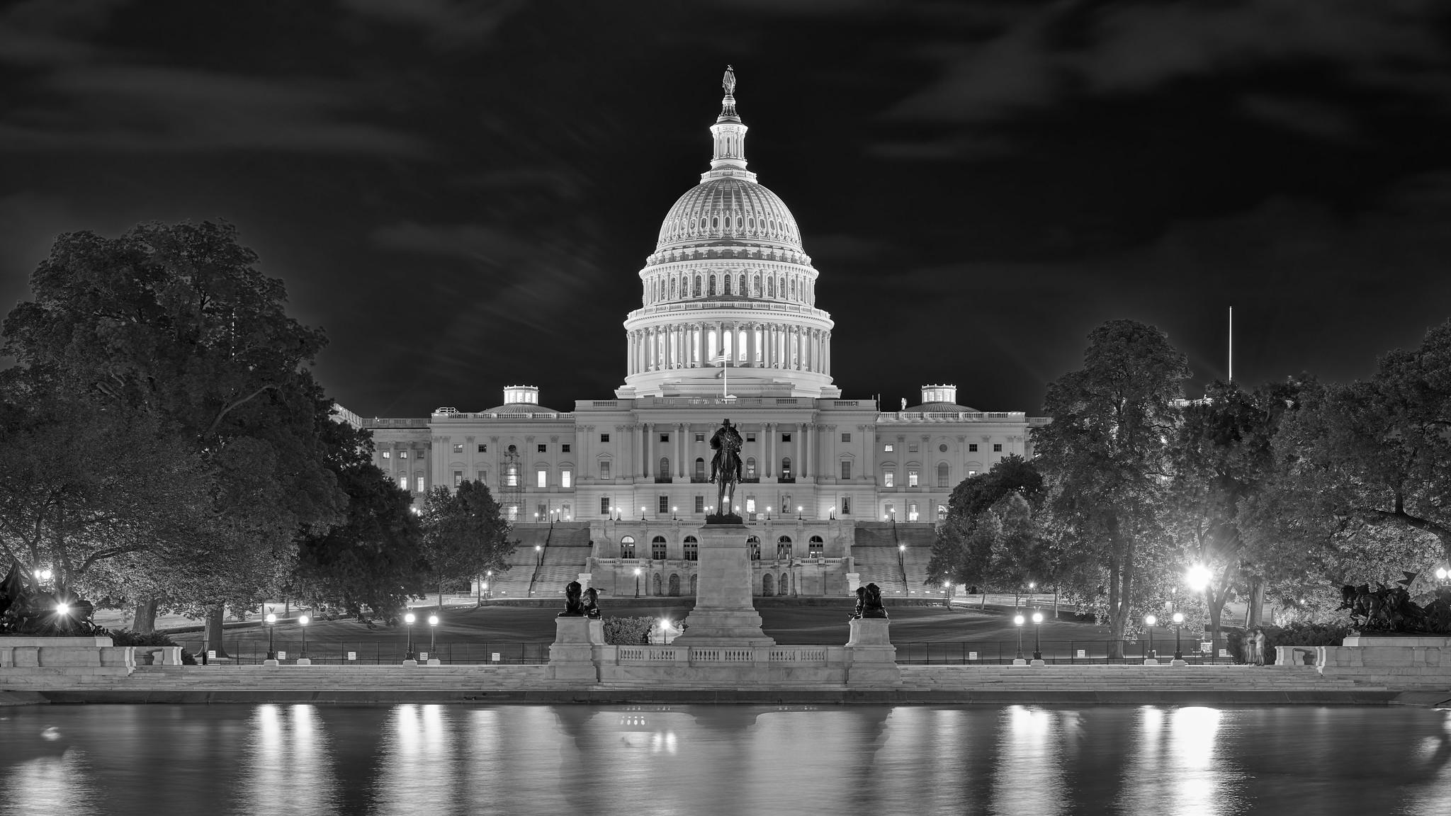 The northern side of the US Capitol building at night, with its prominent dome illuminated. Reflections are visible in the pool at foreground.