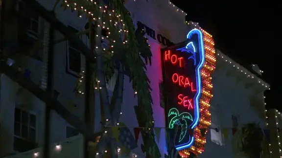 Screenshot from the movie Revenge of the Nerds II showing a neon sign for "Hotel Coral Essex" with strategically burned out letters advertising "Hot oral sex"