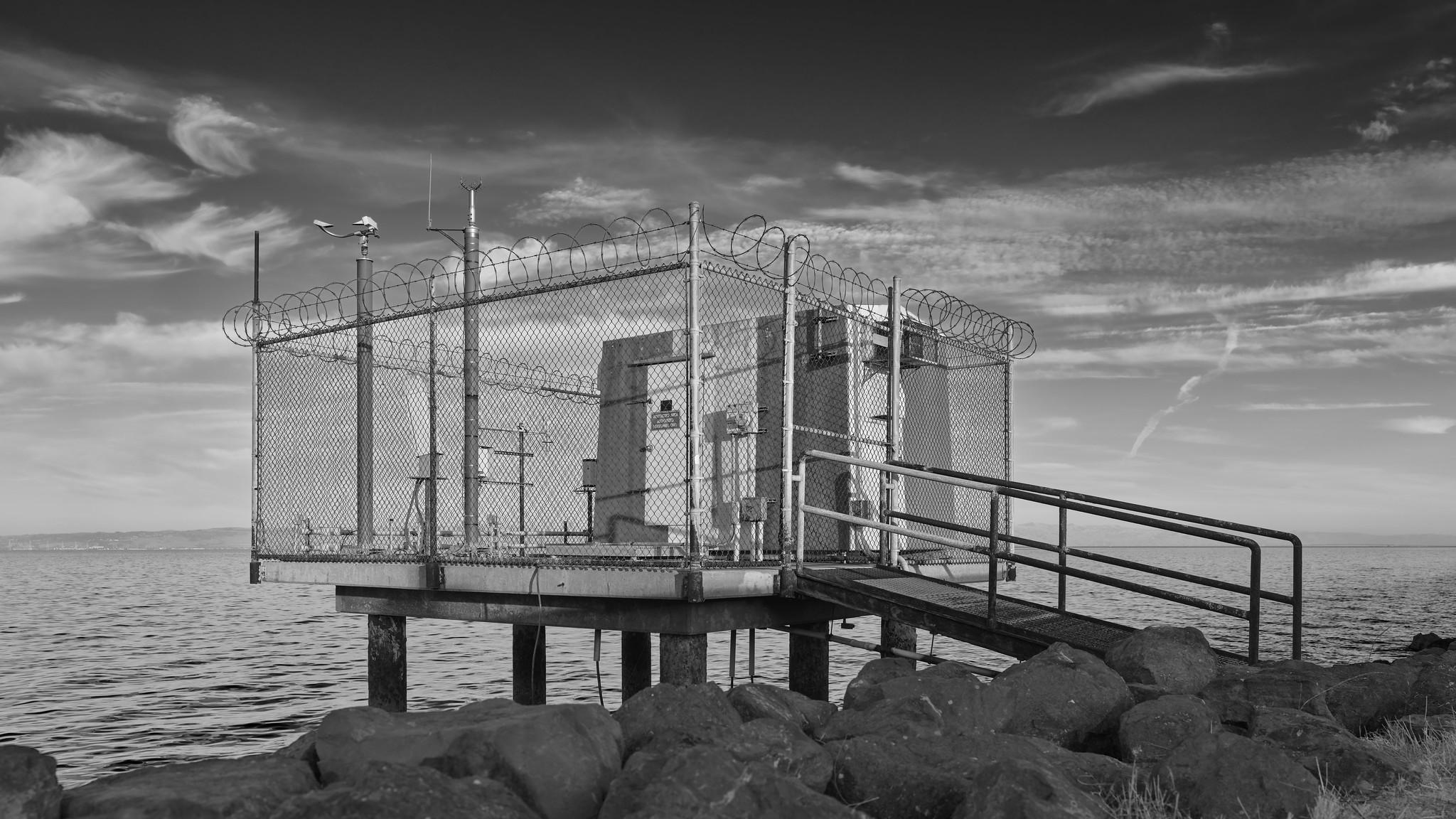 A small shack, surrounded by a chain-link fence and razor wire, with various antennas and masts, on a small pier at the edge of a rocky shoreline.