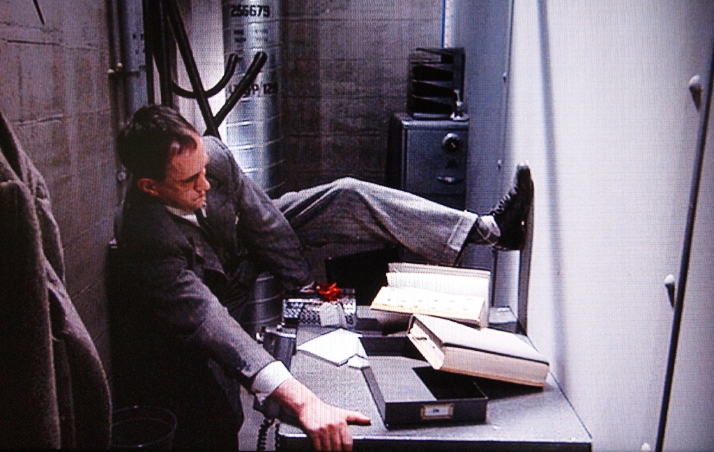 A man in a tiny office split down the middle by a wall, pulling the shared desk more toward his side. From the movie Brazil