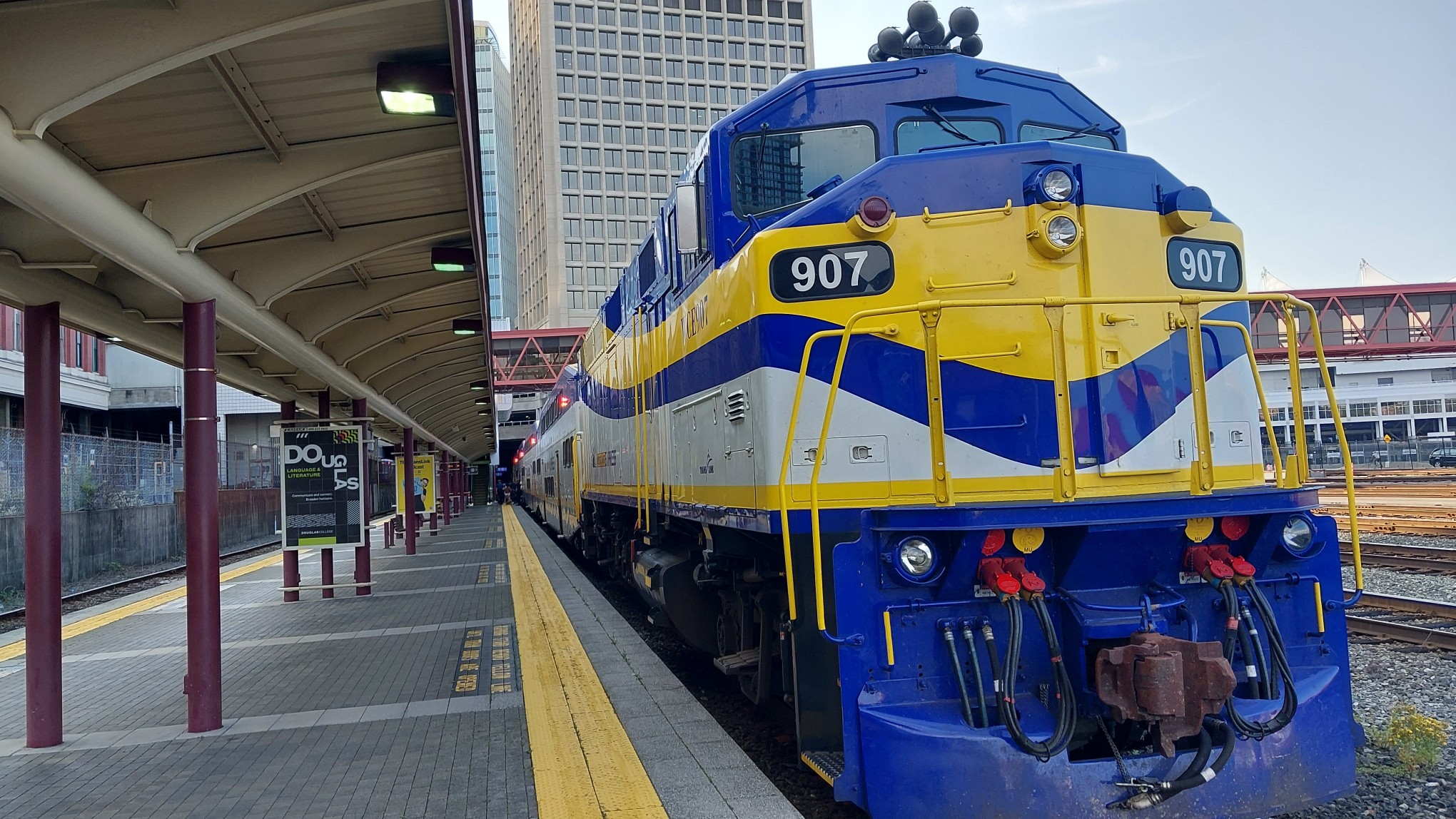 A blue yellow and white train with number plate 907 waits at an empty station platform in downtown Vancouver