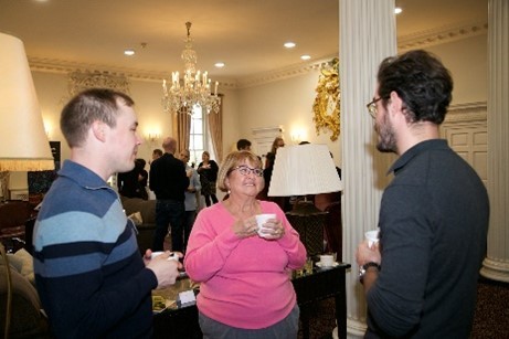 A group of three people in a conference venue engaging with each other gathered in a circle holding a mug, while other scientists gather in the background.