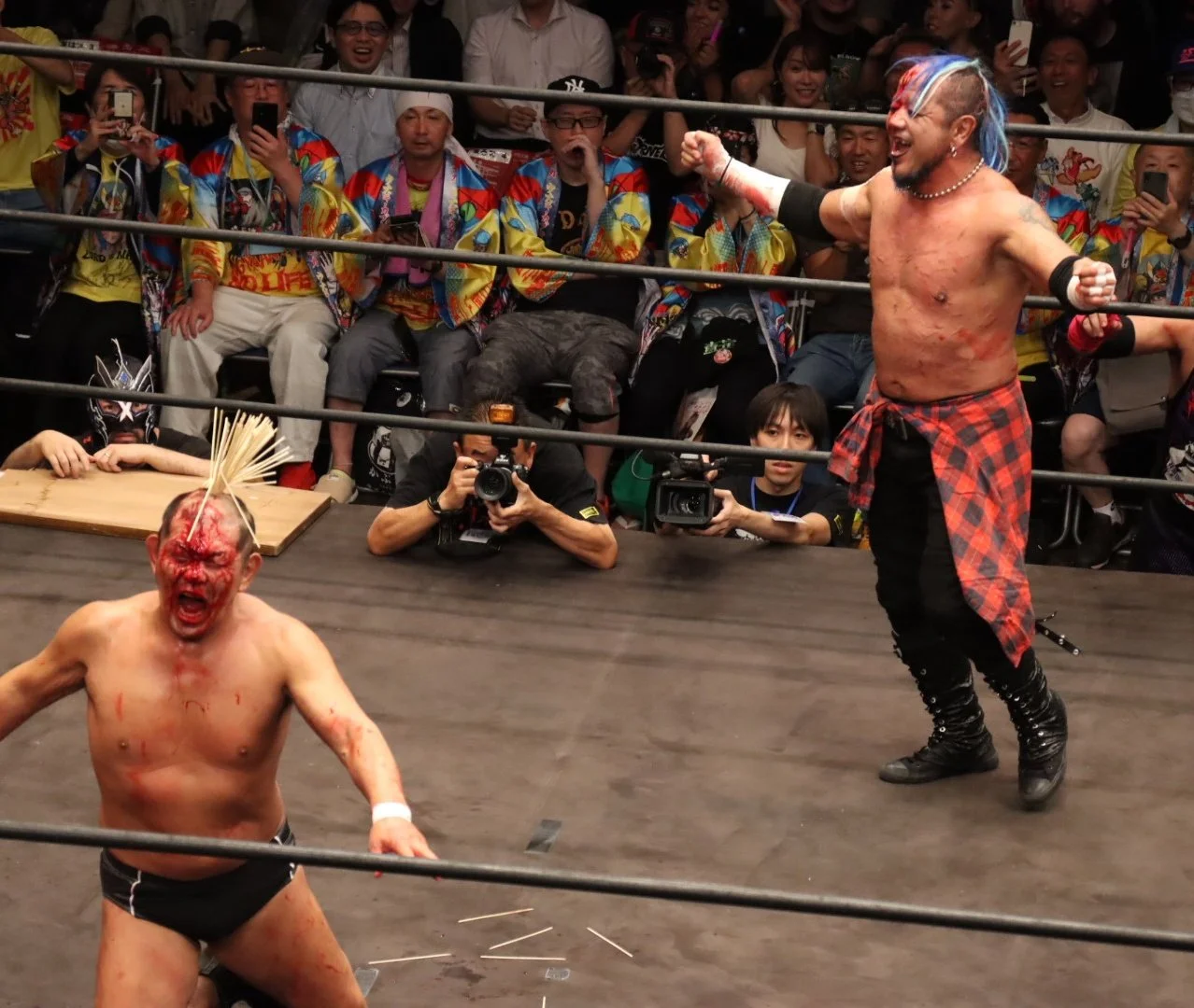 A picture of 2 bloody Japanese pro wrestlers, one with several dozen chopstick/skewers stuck into the top of his head.