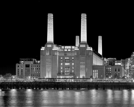 A large industrial building with four prominent smokestacks at each corner, across a river at night.